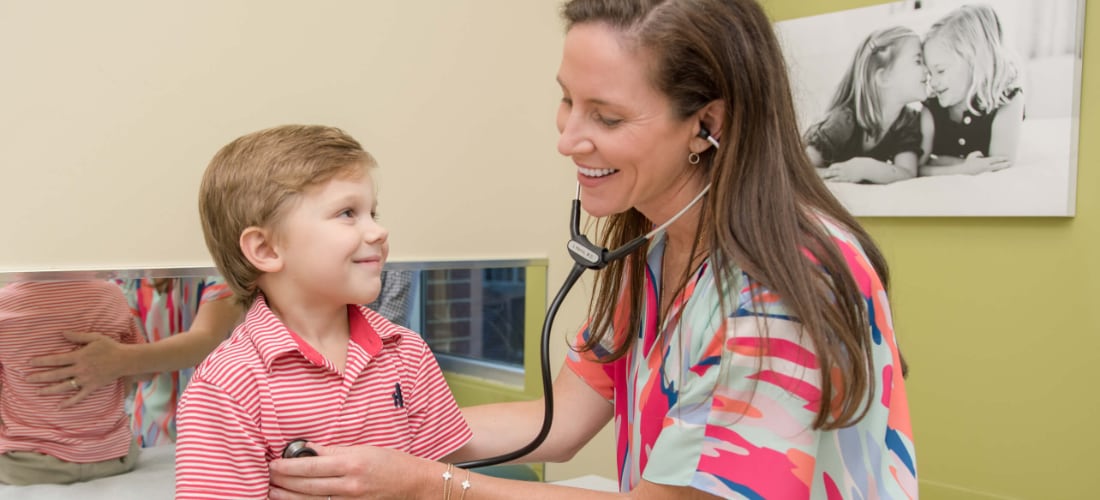 A female doctor smiles as she applies a stethoscope to the chest of a young boy